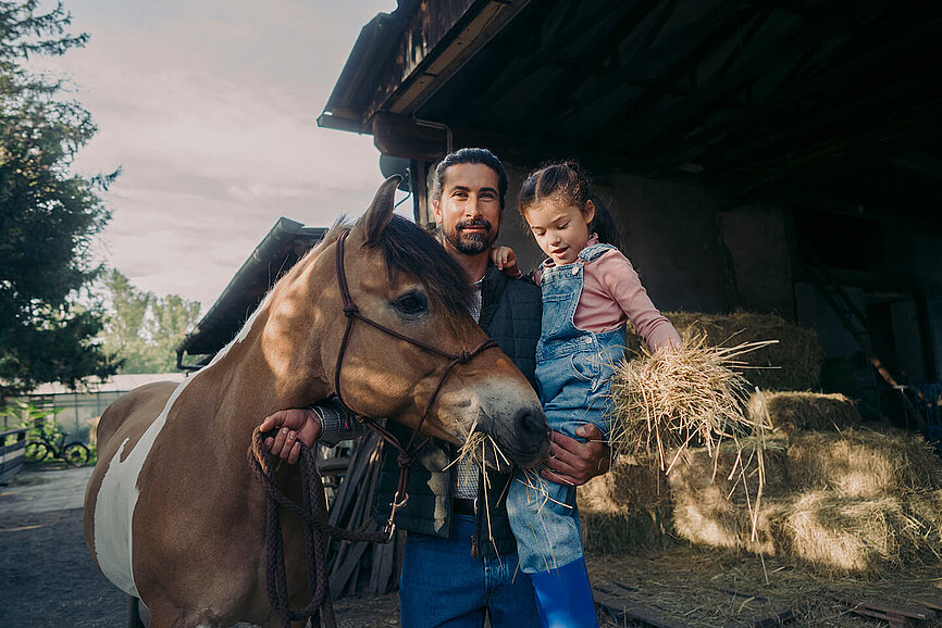 Vater und Tochter stehen im Stall, spielen mit Pferd und Stroh und möchten ihr Vermögen nachhaltig anlegen.