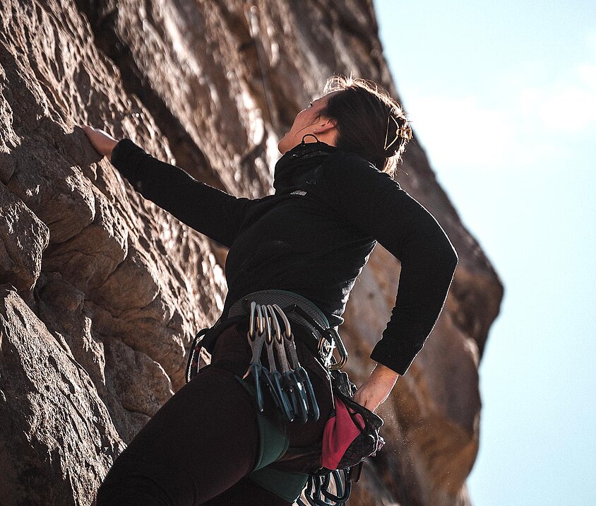 Person in schwarzer Kletterausrüstung erklimmt eine steile Felswand bei Tageslicht.