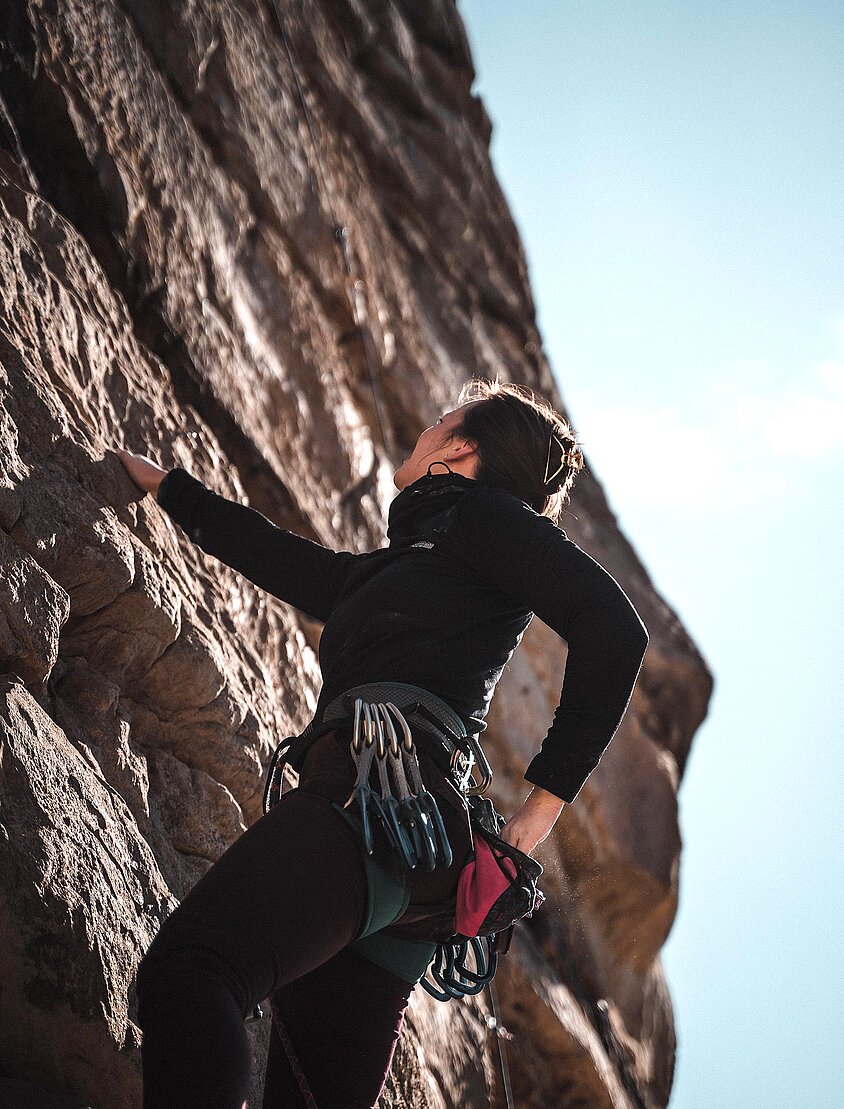 Person in schwarzer Kletterausrüstung erklimmt eine steile Felswand bei Tageslicht.