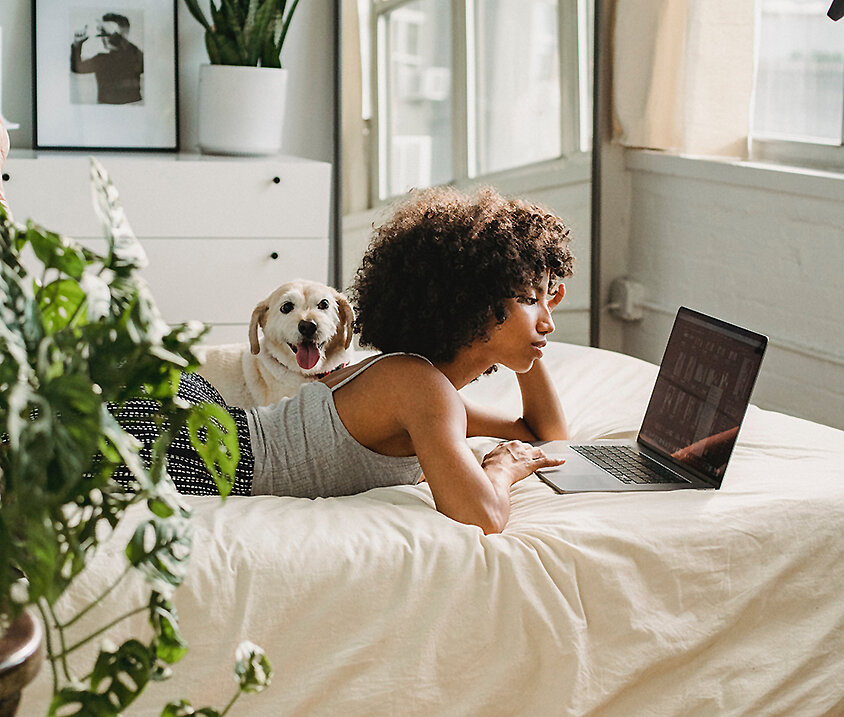Person liegt entspannt auf dem Bett und arbeitet am Laptop in einem hellen Schlafzimmer mit Pflanzen – digitales Leben in sicherem Zuhause.