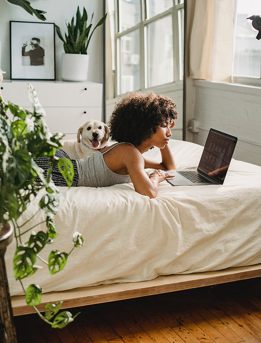 Person liegt entspannt auf dem Bett und arbeitet am Laptop in einem hellen Schlafzimmer mit Pflanzen – digitales Leben in sicherem Zuhause.