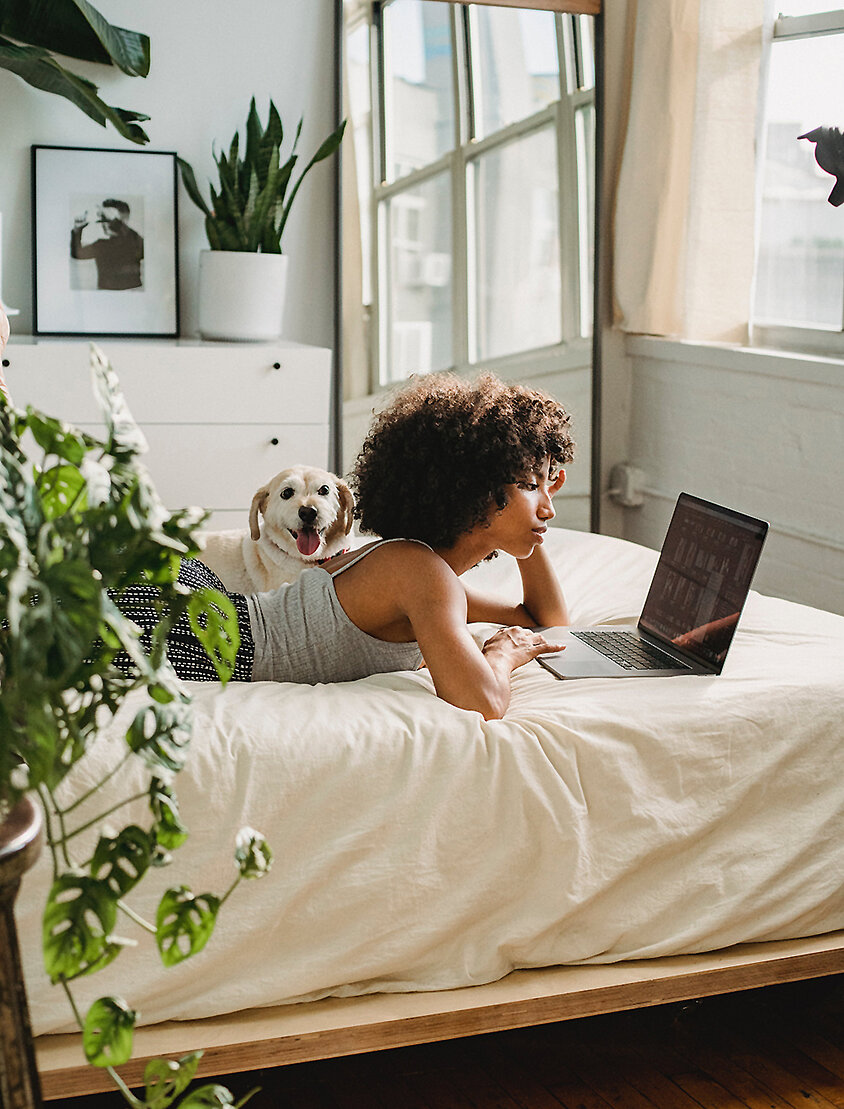 Person liegt entspannt auf dem Bett und arbeitet am Laptop in einem hellen Schlafzimmer mit Pflanzen – digitales Leben in sicherem Zuhause.