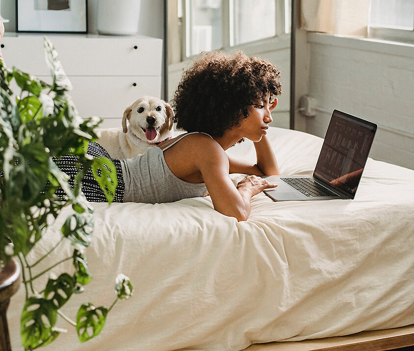 Person liegt entspannt auf dem Bett und arbeitet am Laptop in einem hellen Schlafzimmer mit Pflanzen – digitales Leben in sicherem Zuhause.
