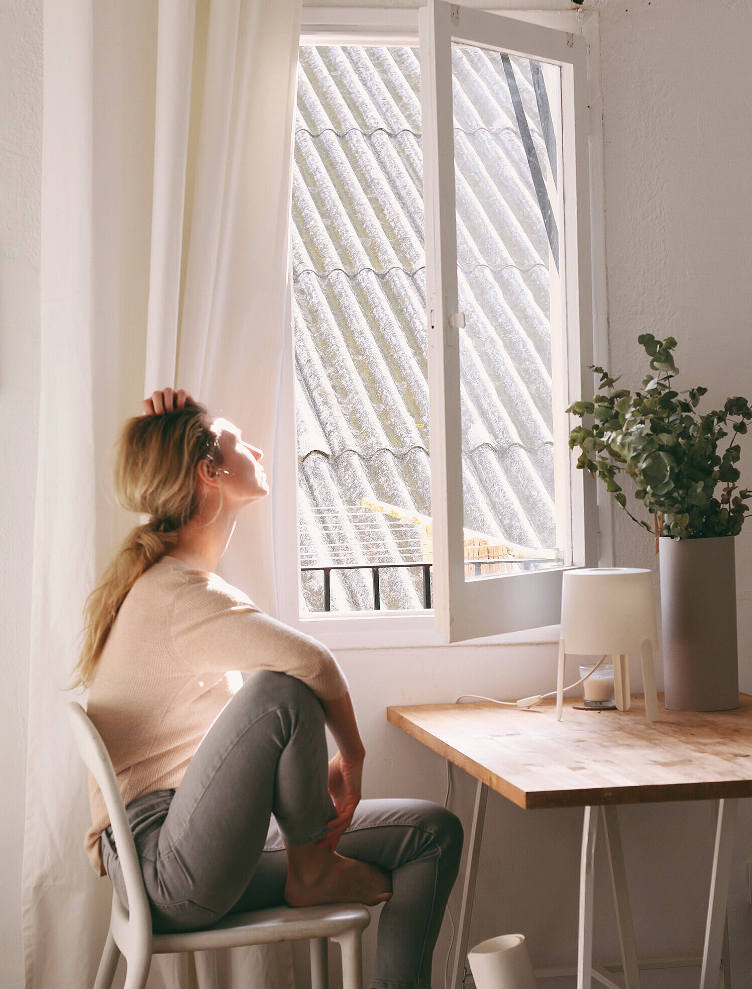 Person sitzt nachdenklich auf einem Stuhl und blickt ins Sonnenlicht durch ein offenes Fenster.