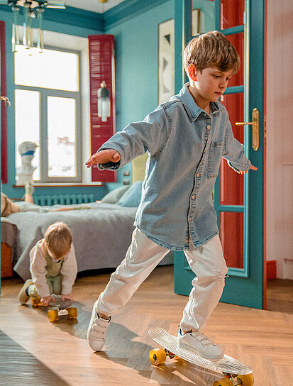 Bruder und Schwester fahren mit dem Skateboard durch die Wohnung und freuen sich, dass ihre Eltern einen Bausparer haben.