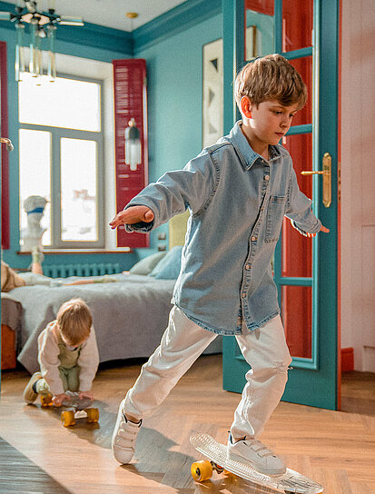 Bruder und Schwester fahren mit dem Skateboard durch die Wohnung und freuen sich, dass ihre Eltern einen Bausparer haben.