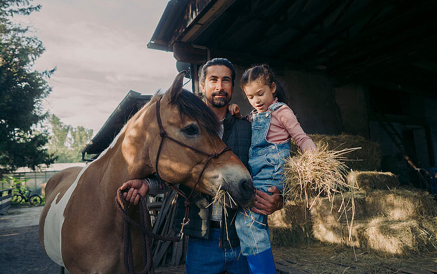 Vater und Tochter stehen im Stall, spielen mit Pferd und Stroh und möchten ihr Vermögen nachhaltig anlegen.