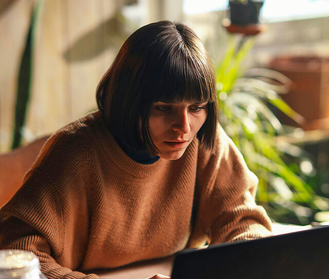 Person im gemütlichen Pullover arbeitet konzentriert an einem Laptop in einer lichtdurchfluteten Wohnung mit Pflanzen. Sie hat schon eine Cyberversicherung abgeschlossen. 