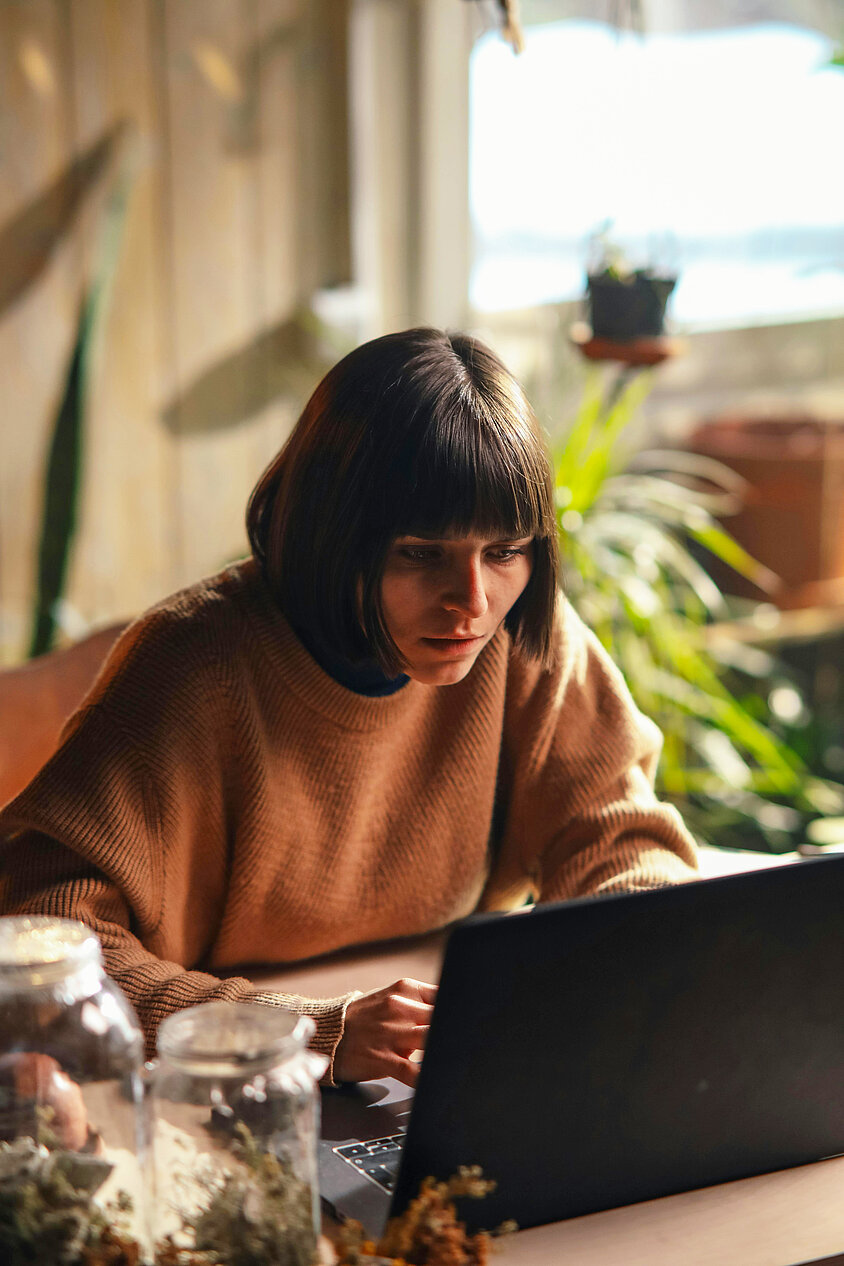 Person im gemütlichen Pullover arbeitet konzentriert an einem Laptop in einer lichtdurchfluteten Wohnung mit Pflanzen. Sie hat schon eine Cyberversicherung abgeschlossen. 