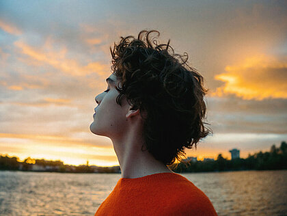 Person mit lockigem Haar im orangen Pullover blickt nachdenklich in den Sonnenuntergang am Wasser und ist froh, eine Unfallversicherung abgeschlossen zu haben. 