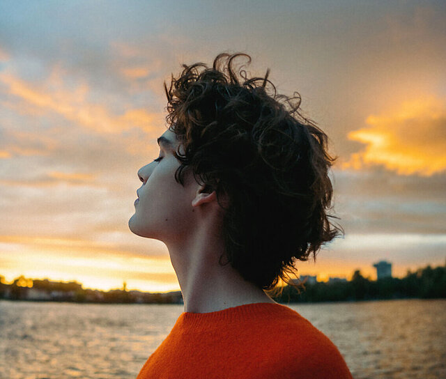 Person mit lockigem Haar im orangen Pullover blickt nachdenklich in den Sonnenuntergang am Wasser und ist froh, eine Unfallversicherung abgeschlossen zu haben. 