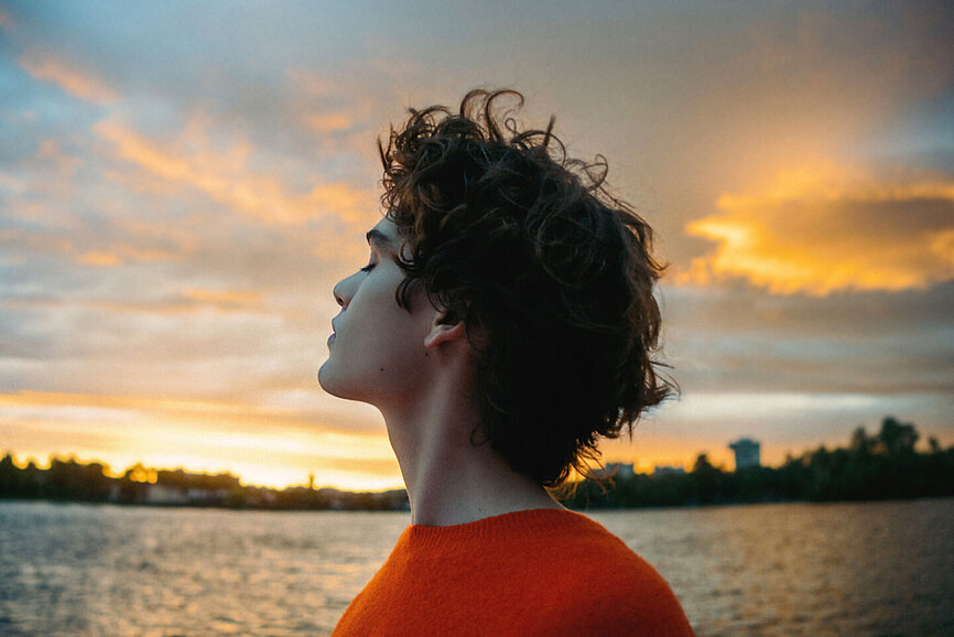 Person mit lockigem Haar im orangen Pullover blickt nachdenklich in den Sonnenuntergang am Wasser und ist froh, eine Unfallversicherung abgeschlossen zu haben. 