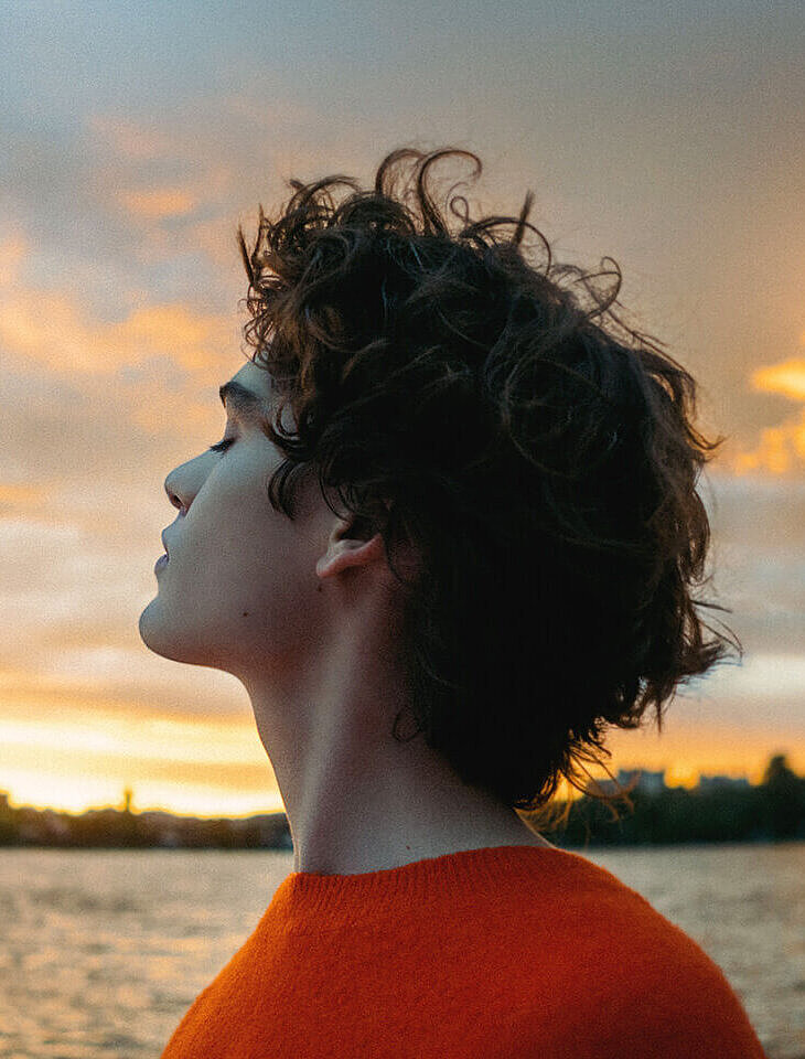 Person mit lockigem Haar im orangen Pullover blickt nachdenklich in den Sonnenuntergang am Wasser und ist froh, eine Unfallversicherung abgeschlossen zu haben. 
