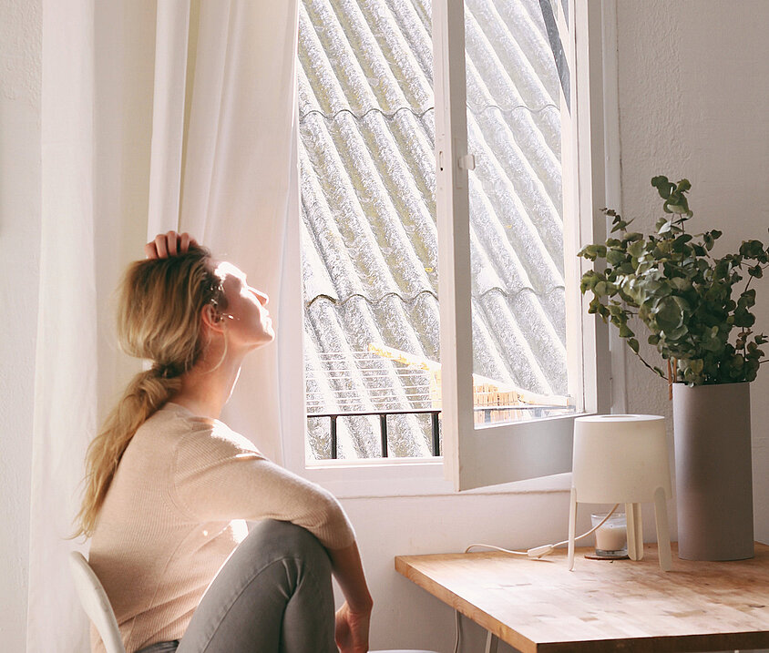 Person sitzt nachdenklich auf einem Stuhl und blickt ins Sonnenlicht durch ein offenes Fenster.