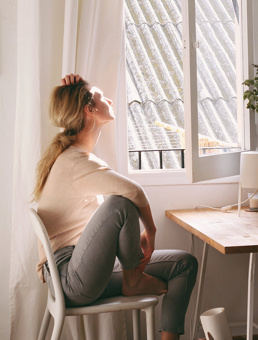 Person sitzt nachdenklich auf einem Stuhl und blickt ins Sonnenlicht durch ein offenes Fenster.