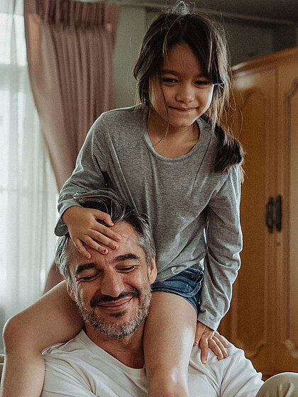 Vater mit Tochter sitzen gemütlich zu Hause im Wohnzimmer und denken über eine Haushaltsversicherung der DONAU nach. 