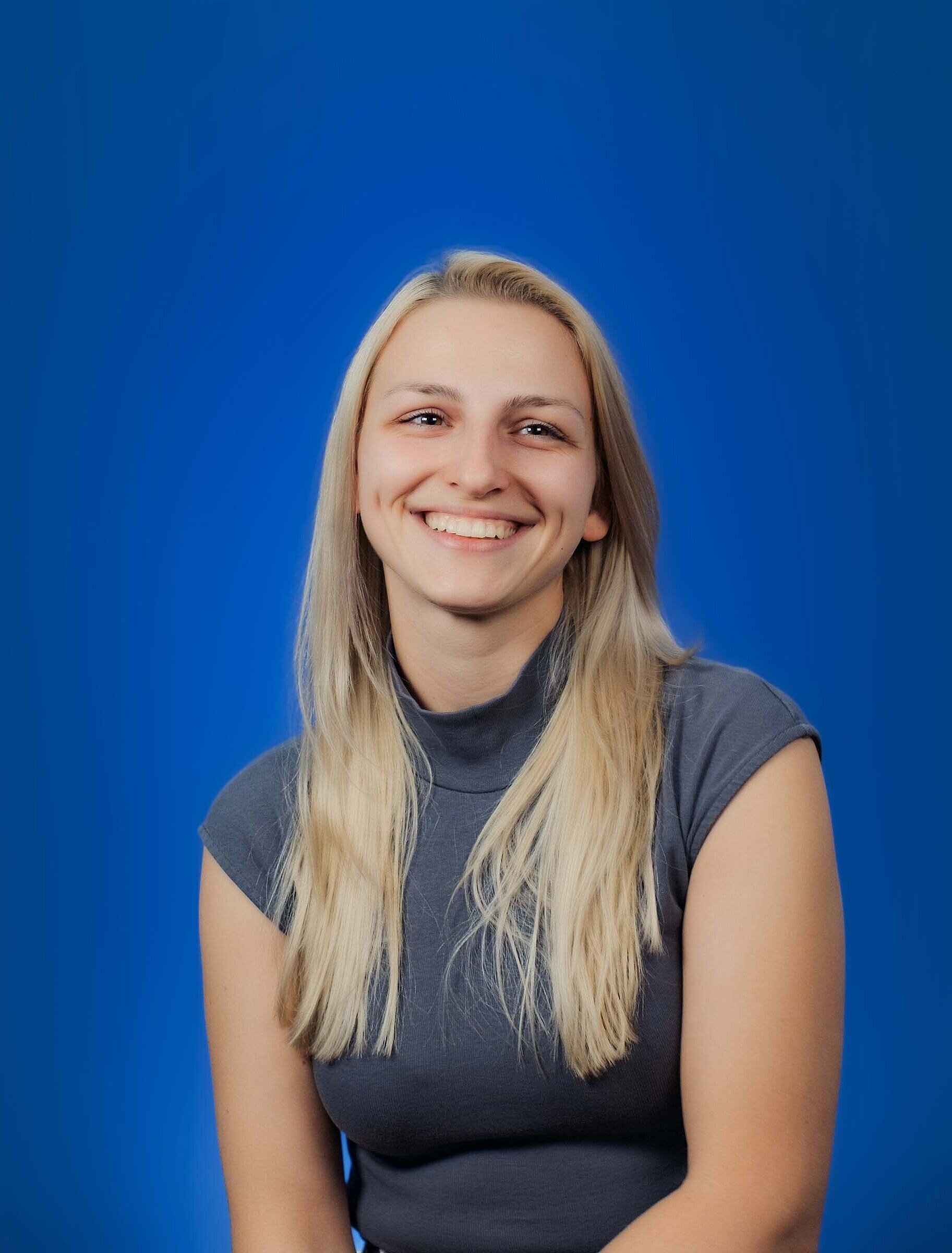 Eine junge Frau sitzt vor einem blauen Hintergrund und lächelt freundlich.
