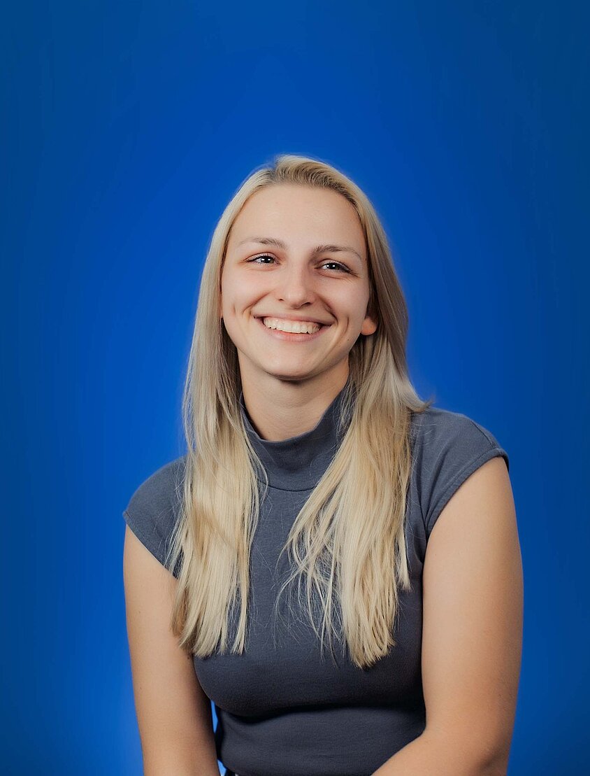 Eine junge Frau sitzt vor einem blauen Hintergrund und lächelt freundlich.