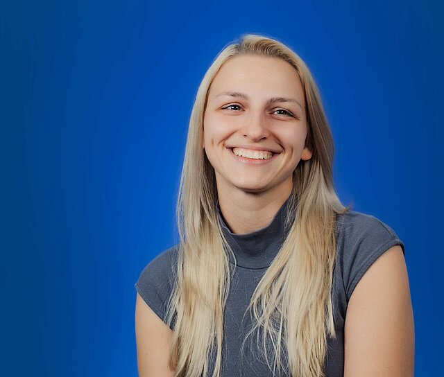 Eine junge Frau sitzt vor einem blauen Hintergrund und lächelt freundlich.