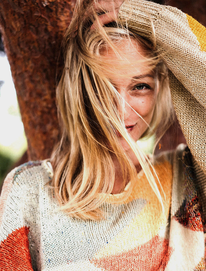 Lächelnde Person mit blondem Haar im bunten Strickpullover, teilweise vom Wind verwehtes Haar im Gesicht.