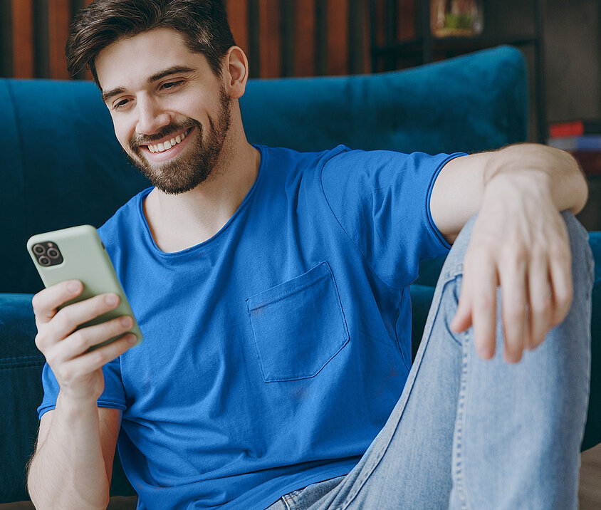 Person mit blauem Shirt sitzt lächelnd vor dem Sofa und informiert sich am Handy über eine Initiativbewerbung bei der DONAU.
