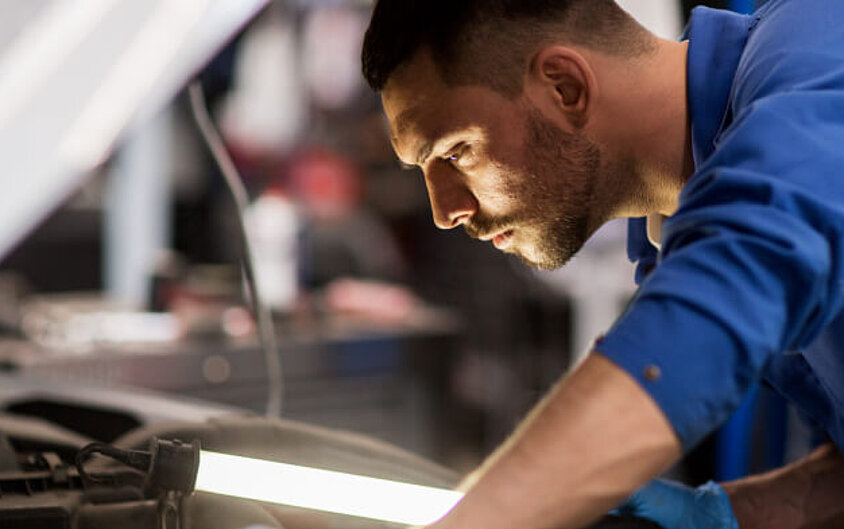 Eine Person in blauem Shirt beugt sich über die geöffnete Motorhaube eines Autos und schaut hinein. Sie ist Mechaniker:in in einer Partner-Werkstätte der DONAU. 